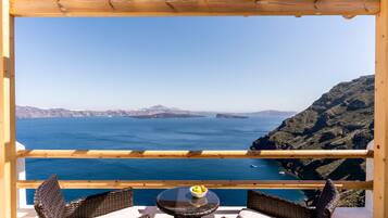 A balcony with wicker furniture and a table with a bowl of fruit, overlooking a coastal landscape with clear blue sea and distant mountains.