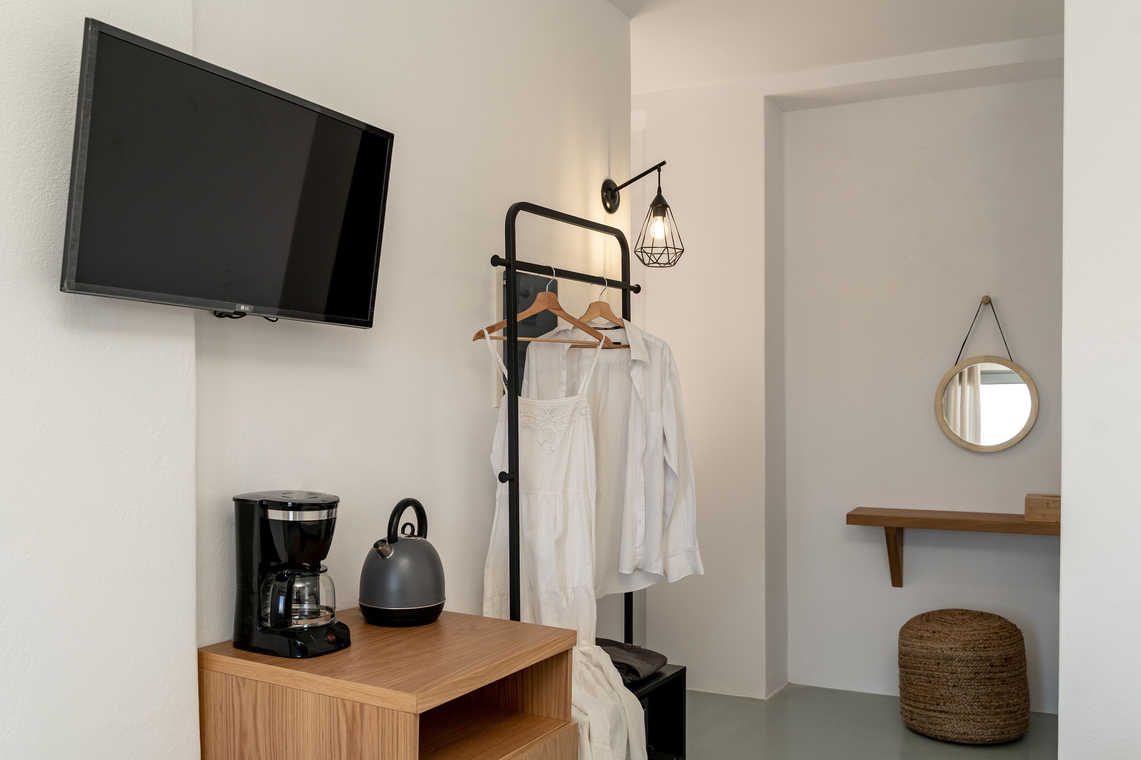 Een moderne kamer met een flatscreen televisie, een koffiezetapparaat, een waterkoker, een kledingrek, een ronde spiegel en een houten plank.