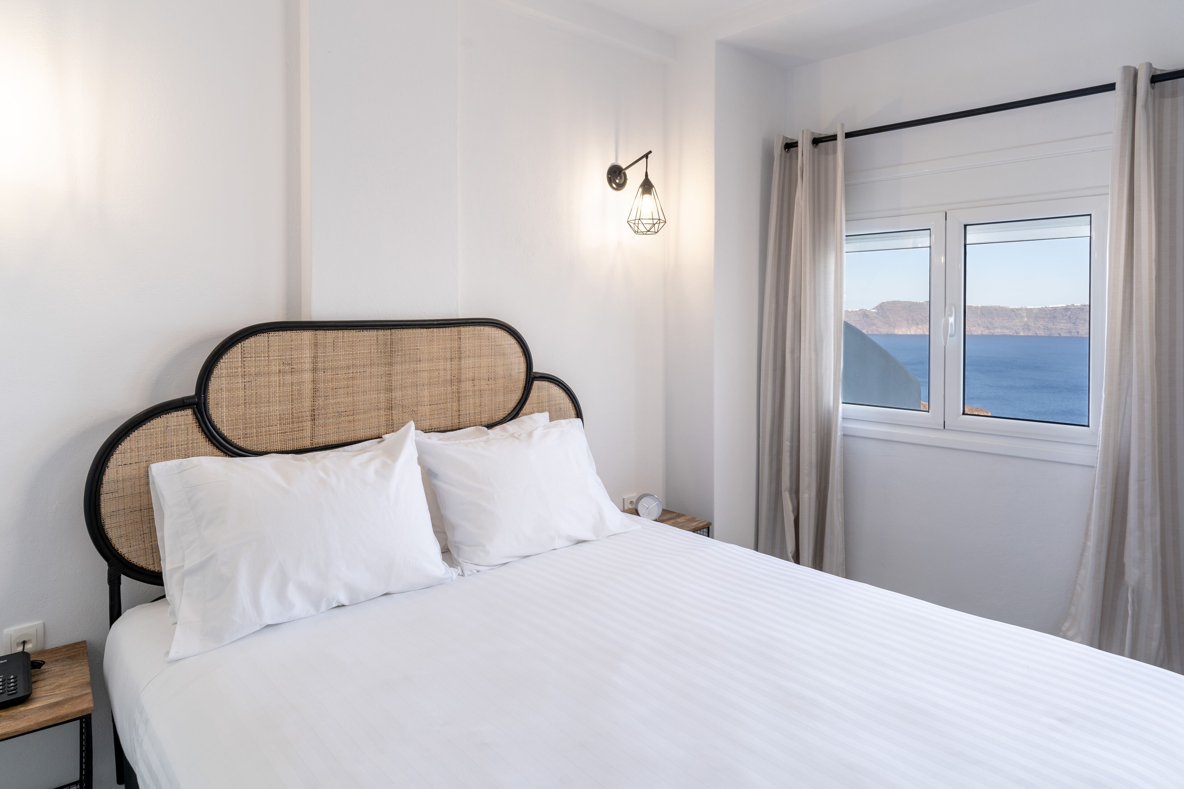 Una cama bien hecha con sábanas blancas, cabecero con diseño tejido, mesita de noche con lámpara y ventana con cortinas.