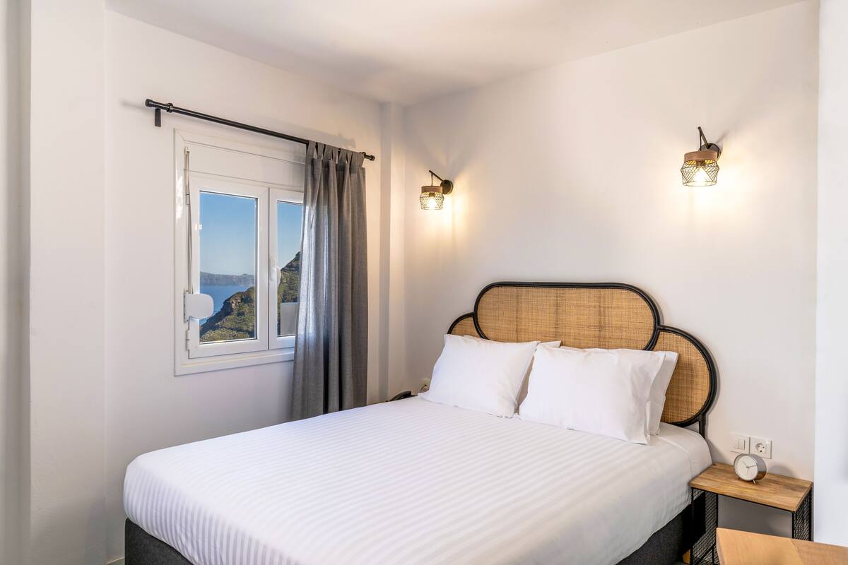 A neatly made bed with white linens, a window with a view of the outdoors, and a bedside table with a clock.