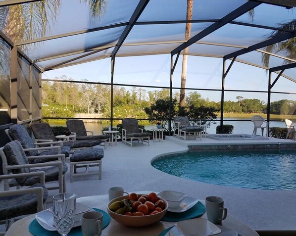 A poolside area with lounge chairs, a table with a bowl of fruit, and a view of a lake and trees.