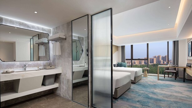Habitación de hotel moderna con una cama grande, un escritorio, una silla, un baño con un espejo amplio y vistas a la ciudad.
