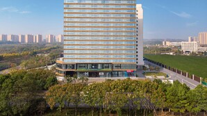 Un moderno edificio de hotel con un llamativo letrero 'MICO', rodeado de vegetación y otros edificios al fondo.