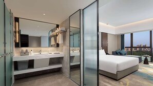 Una habitación de hotel moderna con una cama grande, tocador doble, separador de vidrio y vista a la ciudad.