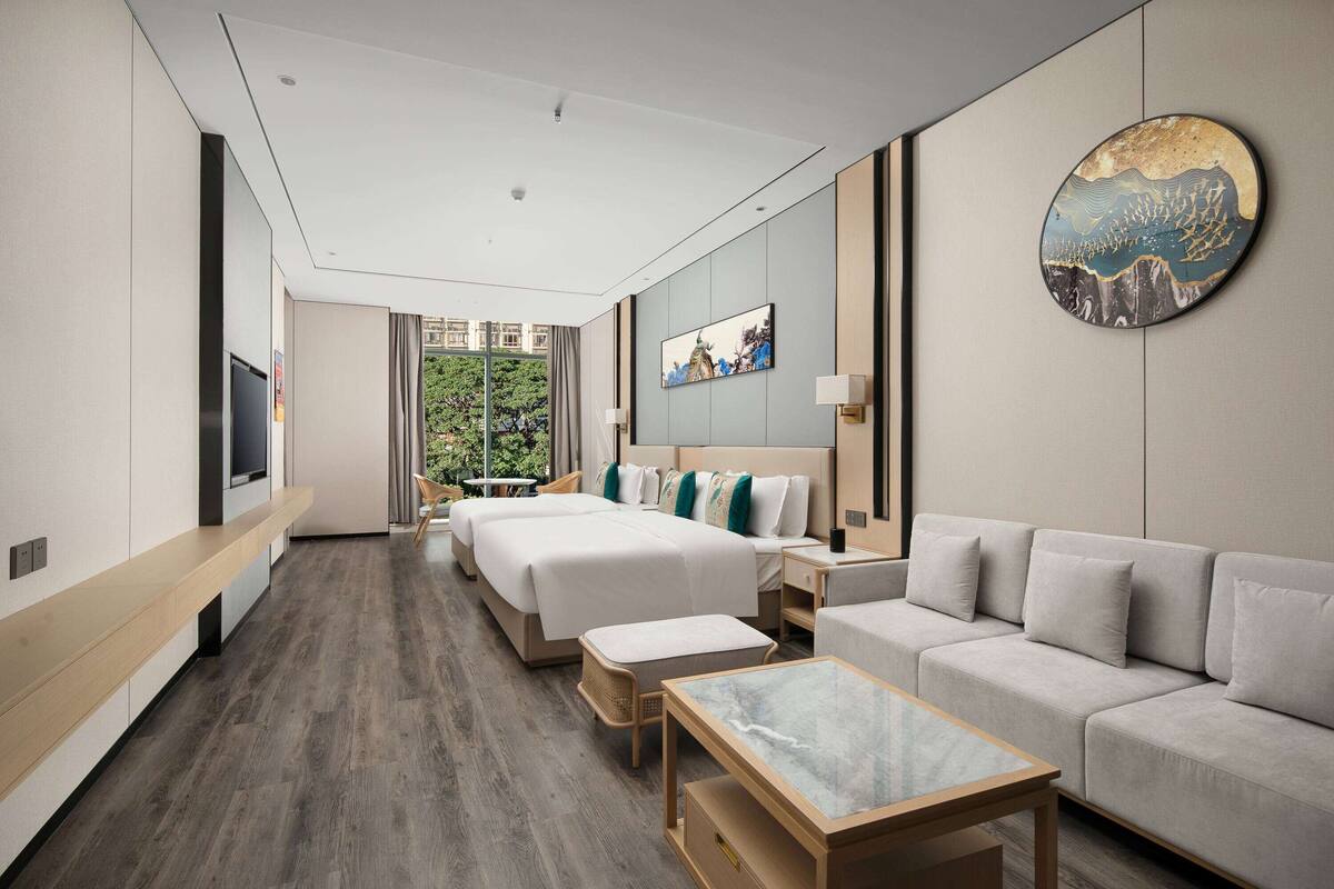 Ein modernes Hotelzimmer mit einem großen Bett, einer Couch, einem Fernseher, einem Esstisch und Blick auf Grünflächen.