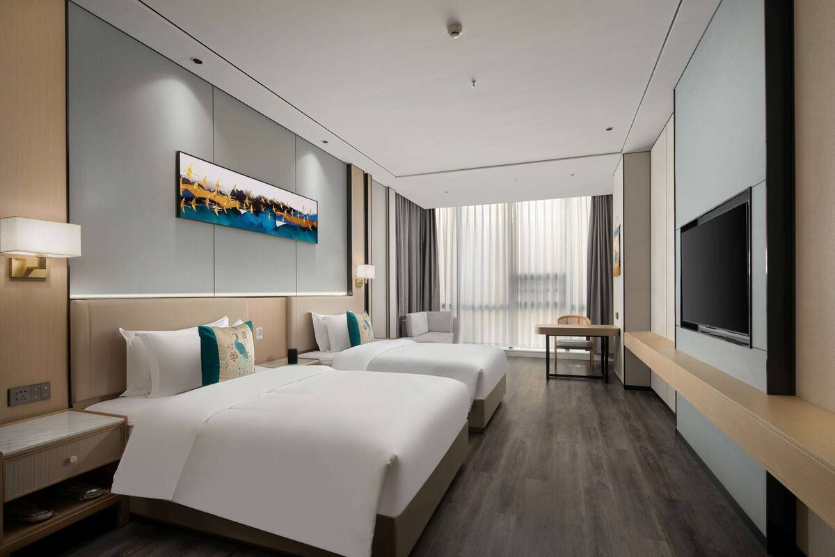 Ein Hotelzimmer mit zwei Betten, einem Flachbildfernseher, einem Schreibtisch und einem großen Fenster mit Vorhängen.