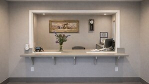 A reception desk with a framed painting above it, a clock on the wall, and a vase with flowers on the desk.