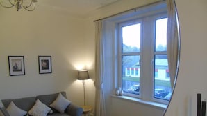 A living room with a grey sofa, a lamp, and a window with curtains. - Snug - Tearlag Apartment (Helensburgh)