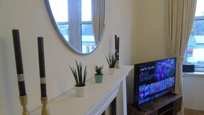 A living room with a fireplace, a TV on a wooden stand, and a window with curtains. - Snug - Tearlag Apartment (Helensburgh)