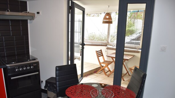 Een keuken met een zwarte kookplaat, een eettafel met rode tafelkleden en uitzicht op het terras door een geopende deur.