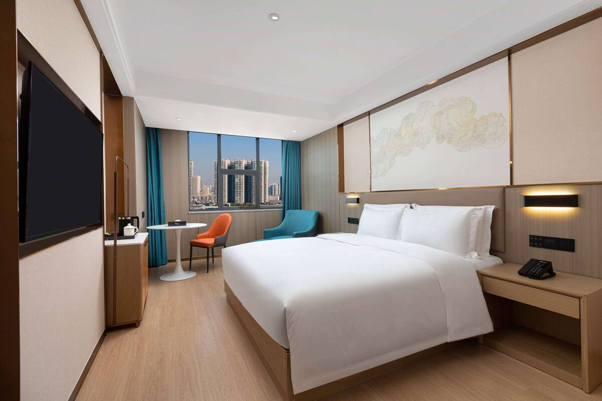 Une chambre d’hôtel avec un grand lit, un bureau avec une chaise, une petite table et une vue sur le panorama de la ville.