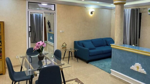 Un salon avec un canapé bleu, une table à manger en verre entourée de chaises, un vase de fleurs et un lustre.