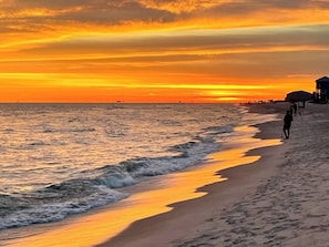 A beach at sunset with waves, a person walking, and a building in the background.