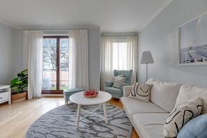 Ein Wohnzimmer mit einer Couch, einem runden Couchtisch, einem Sessel, einer Lampe und einem großen Fenster mit Vorhängen.