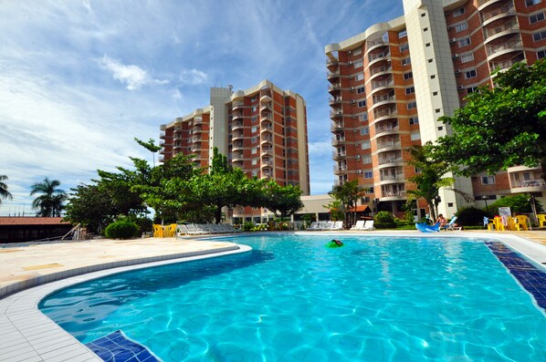 A swimming pool with a curved edge, surrounded by a tiled deck. There are tall residential buildings with balconies in the background.