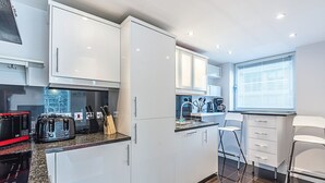 A modern kitchen with white cabinets, a granite countertop, a toaster, a coffee maker, and a window with blinds. - Cosy and comfortable 2 bedroom apartment in Canary Wharf with river view  (London)