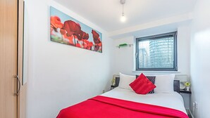 A bedroom with a bed, a window with blinds, a painting on the wall, and a bedside table with a lamp. - Cosy and comfortable 2 bedroom apartment in Canary Wharf with river view  (London)