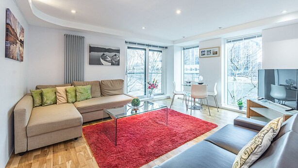 A modern living room with a beige sofa, a red rug, and a TV on a wooden stand. - Cosy and comfortable 2 bedroom apartment in Canary Wharf with river view  (London)