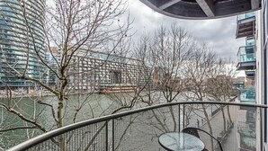 A balcony with a glass table and chairs overlooking a river, surrounded by leafless trees and modern buildings. - Cosy and comfortable 2 bedroom apartment in Canary Wharf with river view  (London)