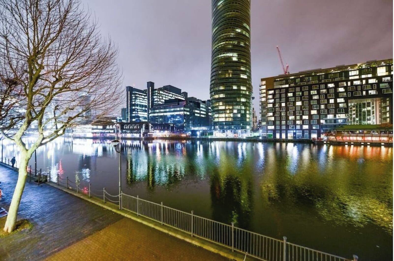 A nighttime view of a cityscape with modern buildings, a river, and a tree-lined walkway.