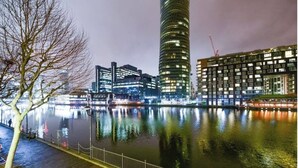 A nighttime view of a cityscape with modern buildings, a river, and a tree-lined walkway. - Cosy and comfortable 2 bedroom apartment in Canary Wharf with river view  (London)