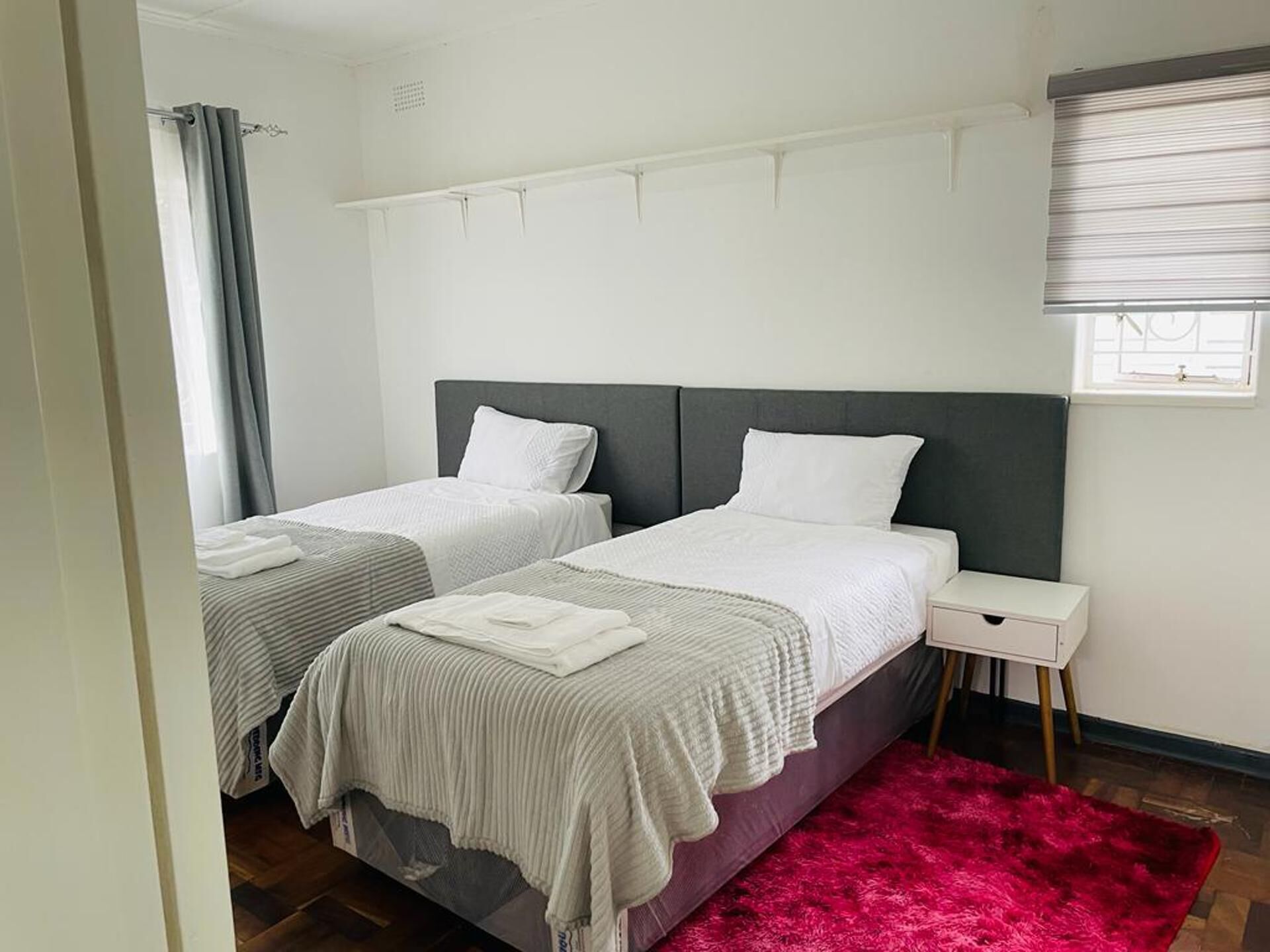 Habitación de hotel con dos camas individuales, una mesita de noche pequeña, una ventana con persianas y una alfombra roja.