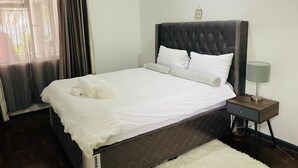 Un lit bien fait, avec du linge de lit blanc et des oreillers, une tête de lit sombre, une table de chevet avec une lampe et une fenêtre avec des rideaux.