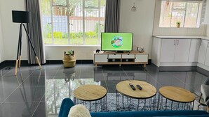 Un salon moderne avec un canapé bleu, un téléviseur posé sur un meuble en bois et un coin cuisine doté de meubles blancs.