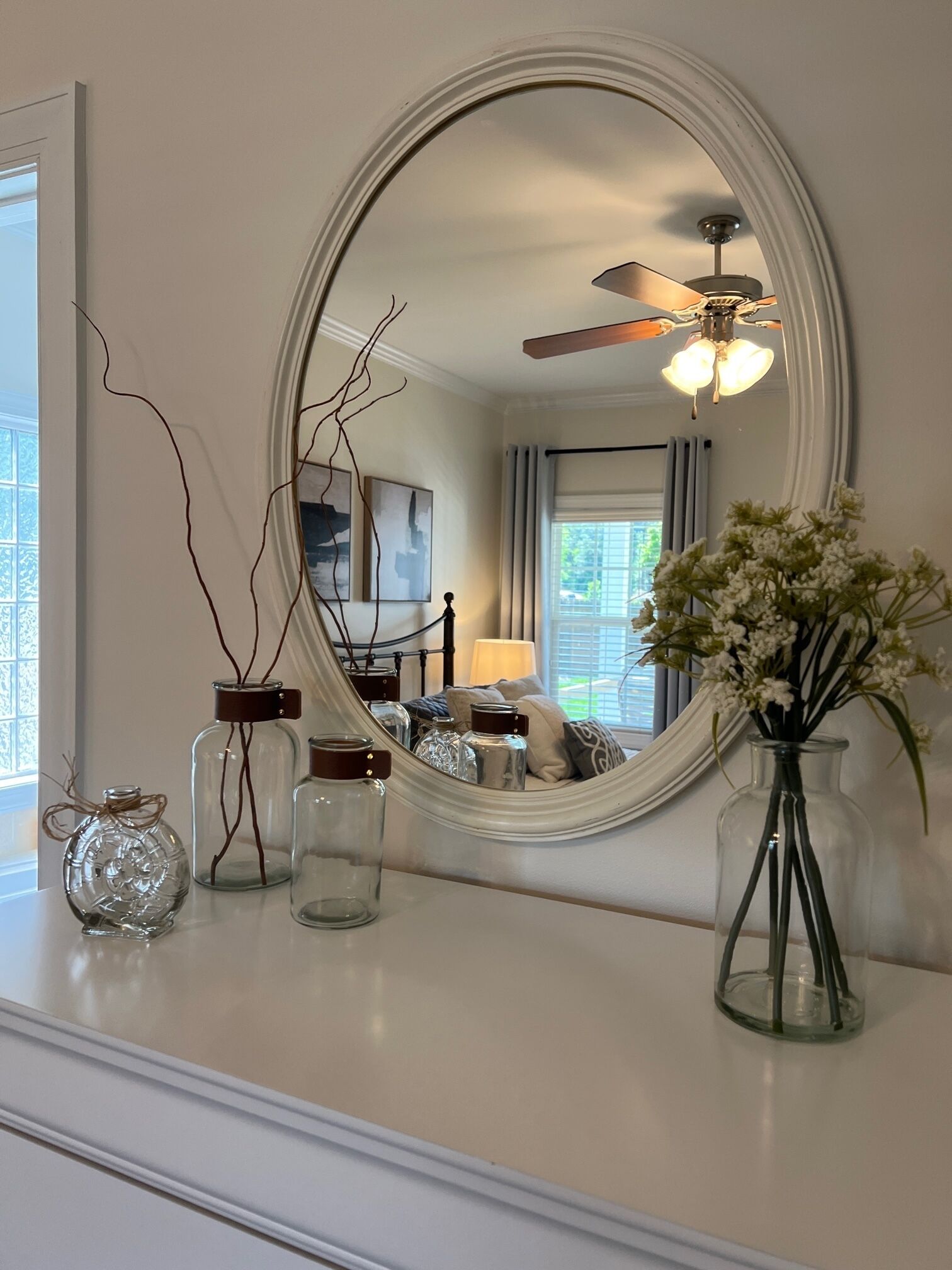 A room with a large oval mirror, a ceiling fan, and a window with curtains.