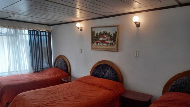 A hotel room with three beds, each with a wooden headboard and orange bedding. There is a painting on the wall and a window with curtains. - GH Plaza (Riobamba)