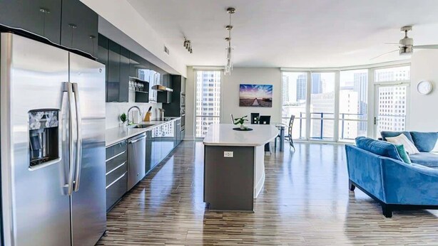 Cozinha moderna com ilha central, eletrodomésticos de aço inoxidável e um sofá azul numa sala de estar luminosa com grandes janelas.