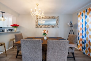 A dining area with a wooden table, four grey upholstered chairs, a chandelier, a mirror with an ornate frame, and a vase with flowers.
