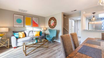 A modern living room with a sofa, coffee table, and chairs. There is a dining table with placemats. The room has artwork on the walls and a chandelier.