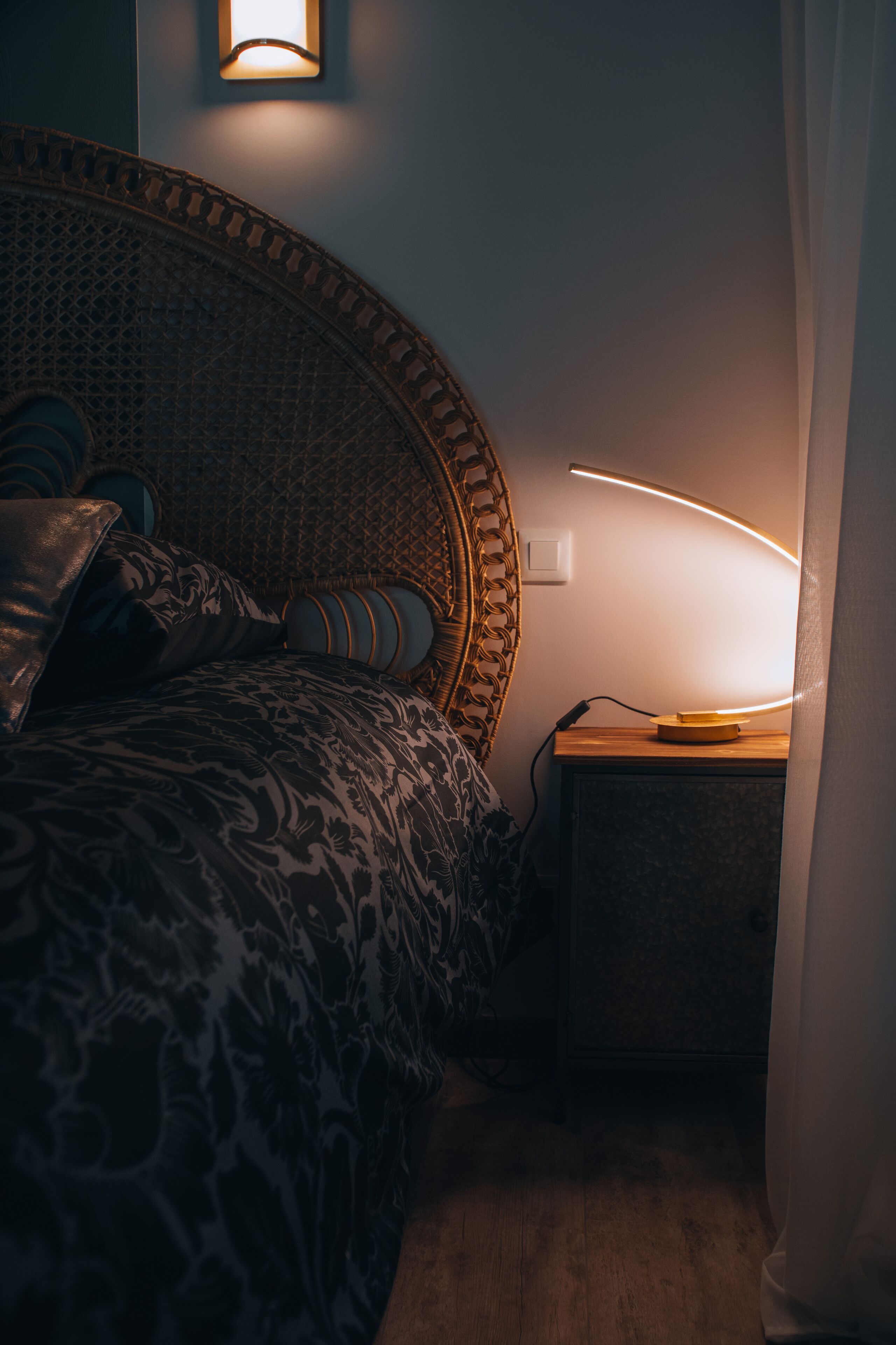 Een slaapkamer met een bed met een patroon, een gebogen hoofdeinde, een bedlampje en een stopcontact aan de muur.