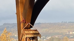 Un balcó de fusta amb un allotjament per ocells i una cadira, amb vista a un paisatge rural de camps i cases.