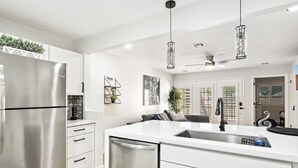 Uma cozinha moderna com geladeira de aço inoxidável, armários brancos, uma ilha central com pia, luminárias pendentes e um ventilador de teto.