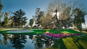 Um campo de golfe com um lago refletindo árvores e um gramado bem cuidado com flores coloridas.
