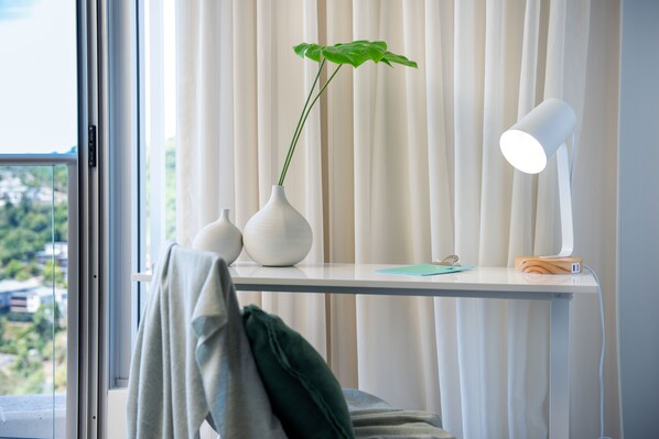 Quarto com escrivaninha branca, planta em vaso, luminária e vista para o verde lá fora.