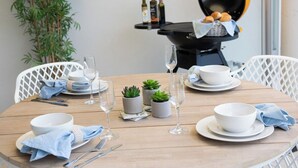 Et rundt spisebord med hvide tallerkener, skåle og glas, ledsaget af blå servietter og bestik. Der er små krukkeplanter og en grill i baggrunden.
