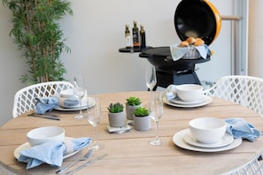 A round dining table with white plates, bowls, and glasses, accompanied by blue napkins and silverware. There are small potted plants and a grill in the background. - Pacific Blue Whitsunday - 2 Bedroom (Airlie Beach)