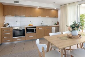 A modern kitchen with wooden cabinets, a built-in oven, a microwave, and a dining area with a table and chairs. - Pacific Blue Whitsunday - 2 Bedroom (Airlie Beach)