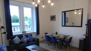 A living room with a sofa set, a dining table with chairs, a large mirror, and a window with curtains. - Snug - Sealladh Mara Apartment (Helensburgh)