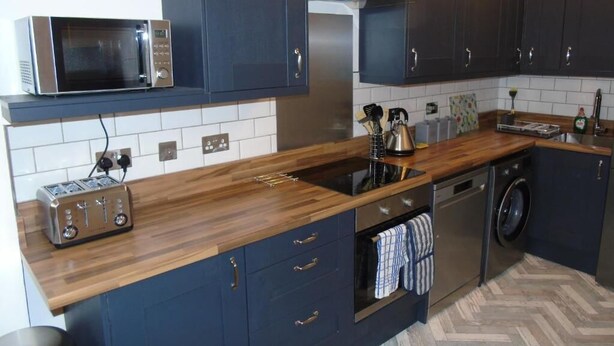 A modern kitchen with dark cabinets, a microwave, a toaster, a sink, and a washing machine. - Snug - Sealladh Mara Apartment (Helensburgh)