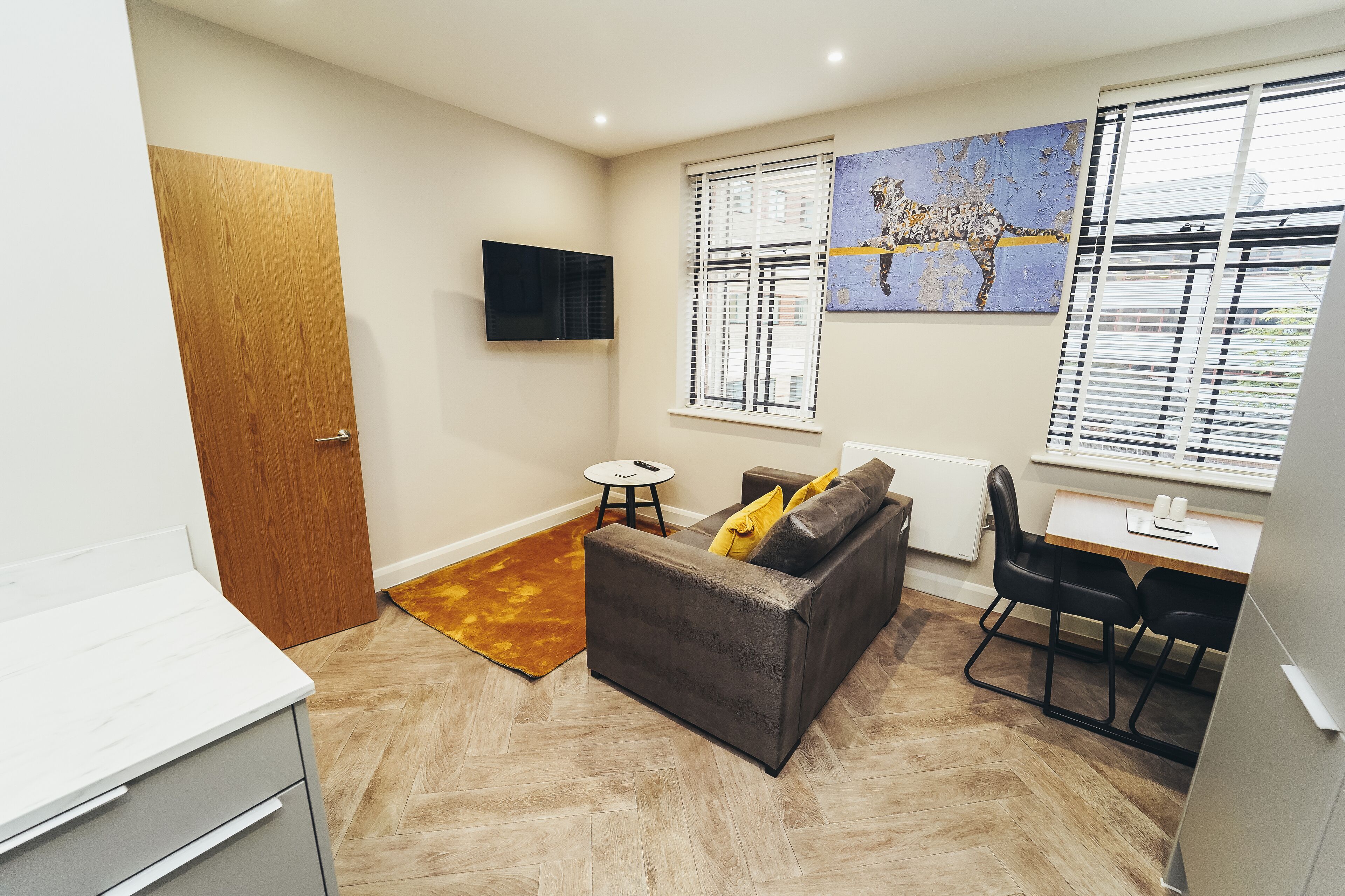 A compact living space with a sofa, a small table, a TV mounted on the wall, and a window with blinds.