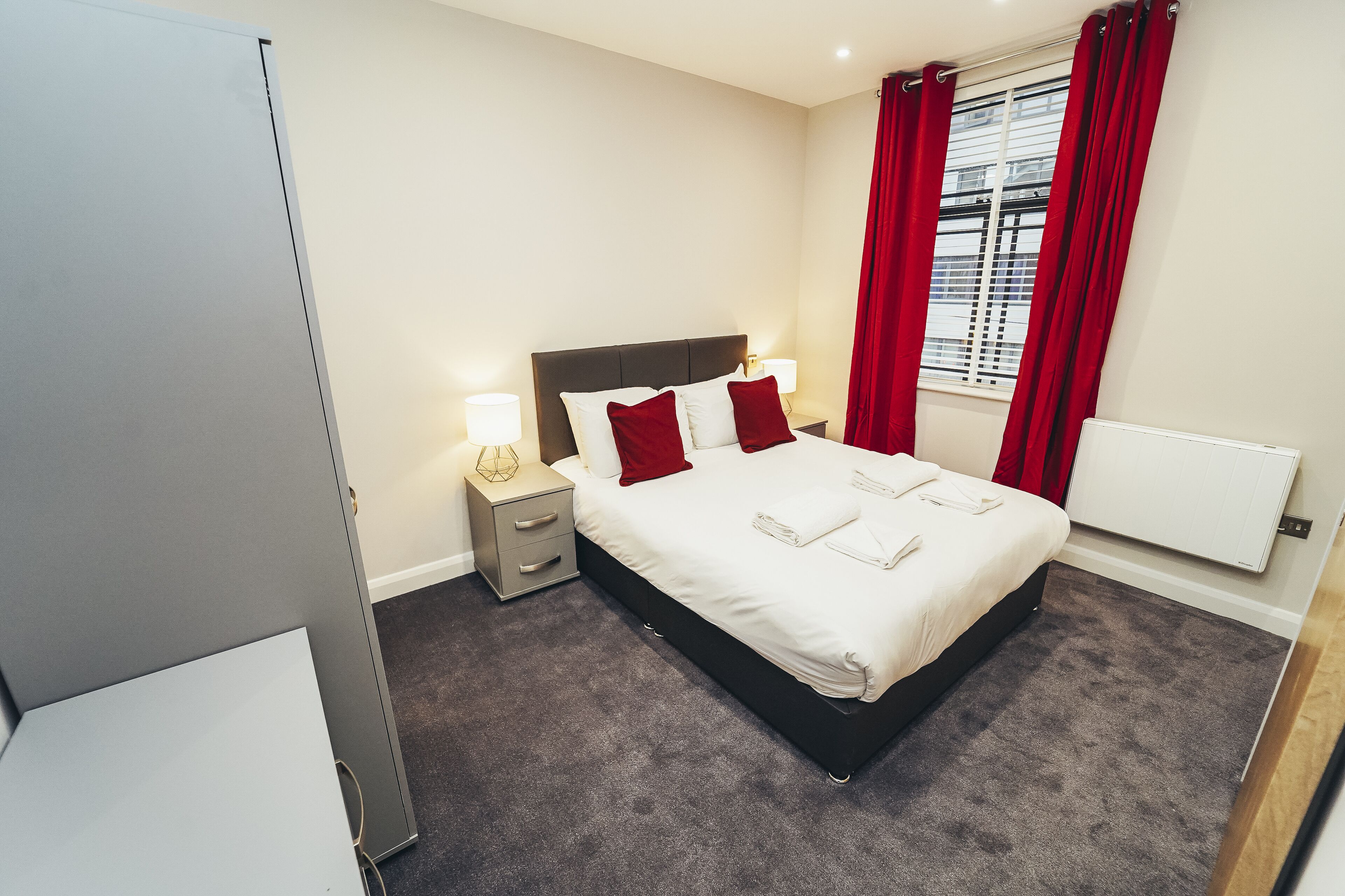 Un dormitorio ordenado con cama, lámparas de noche y una ventana con cortinas rojas.