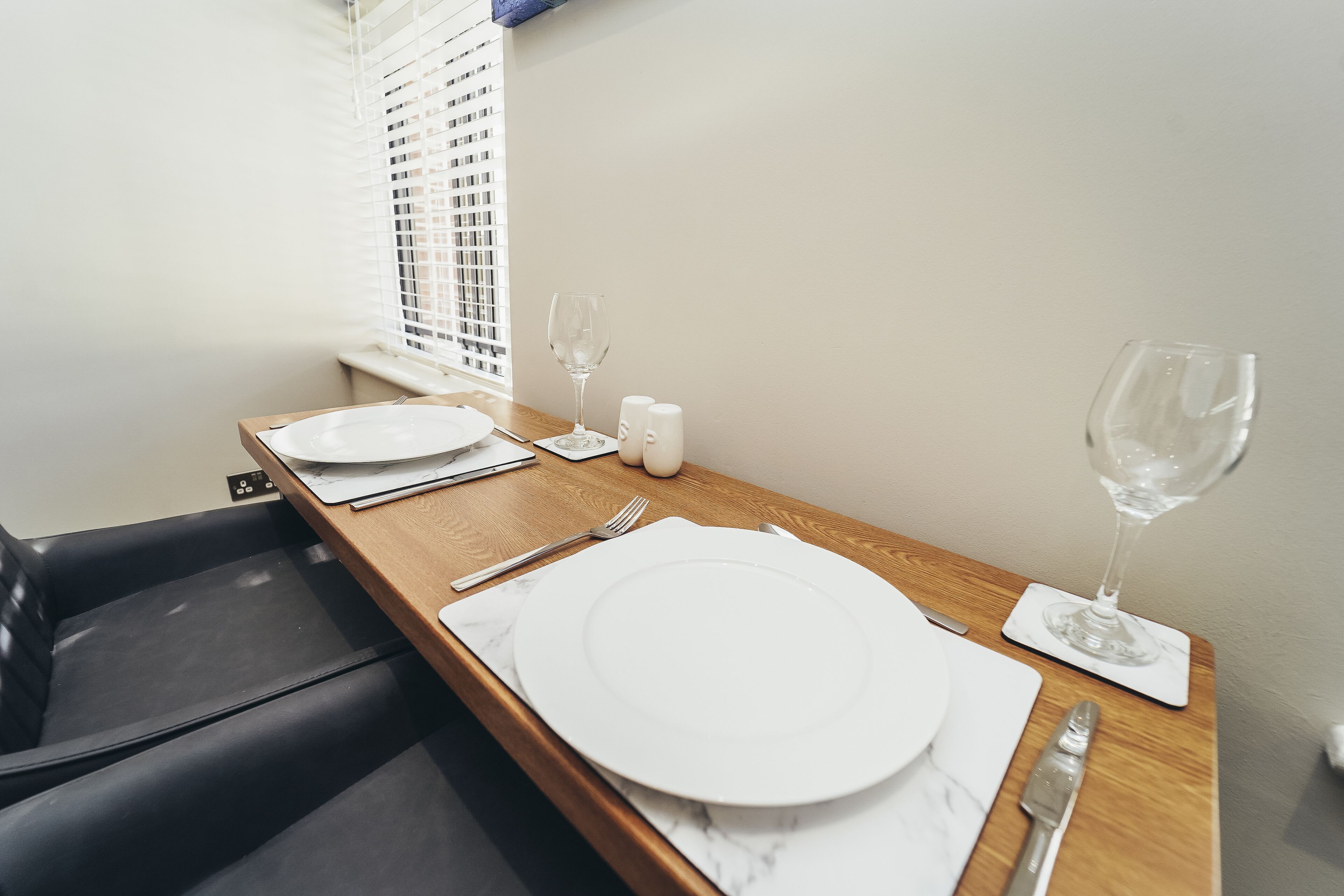 Una mesa de comedor dispuesta con platos blancos, cubiertos y copas de vino, con una ventana y persianas al fondo.