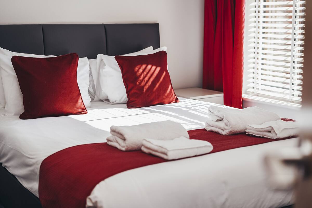 A neatly made bed with white and red bedding, a dark headboard, and a window with blinds.