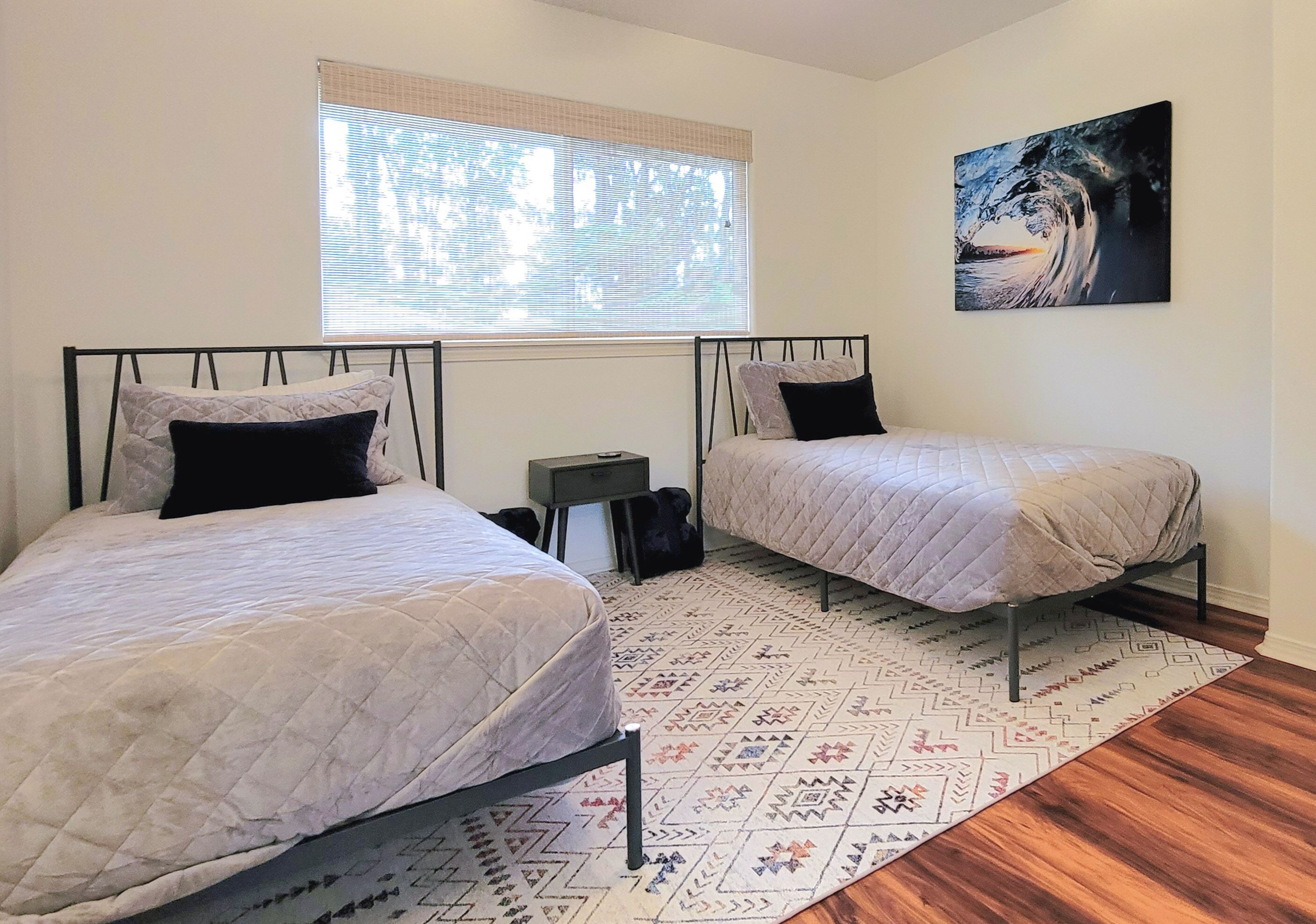 Un dormitorio con dos camas, una ventana con persianas, un cuadro en la pared y una alfombra estampada.