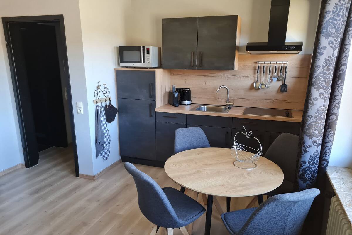 Een kleine keuken met een magnetron, gootsteen en kastjes. Er staat een rond tafel met twee stoelen en een decoratief object erop.