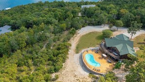 An aerial view of a resort with a swimming pool, a large building with a green roof, and dense forest surrounding the property. - 5BR Table Rock Lake Getaway |Hot Tub, Pool Games (Blue eye)